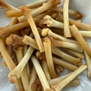 May include: A pile of light tan, bone-shaped dog treats with darker, textured ends. The treats are scattered, showing their individual shapes and sizes. The image is taken from a close-up perspective, highlighting the details of the treats.