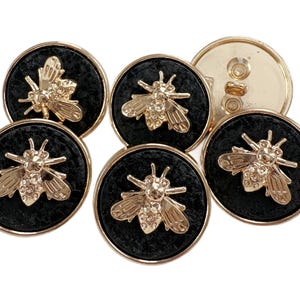 May include: Set of six decorative buttons. Each button features a gold-colored bee design on a black, textured background. The buttons have a gold-colored rim and a gold-colored back with two holes for sewing.