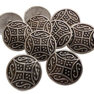 May include: A collection of silver-toned metal buttons with an ornate, symmetrical design. Each button features a central square pattern with dotted details and swirling embellishments. The buttons are round and have a vintage aesthetic.