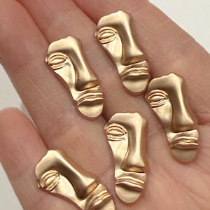 May include: Six gold-colored face-shaped charms with detailed features, including eyes, nose, and lips. The charms are arranged on a light background, showcasing their metallic finish and artistic design. These charms are ideal for jewelry making.