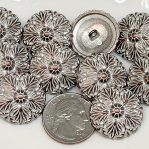 Op de afbeelding: Een verzameling zilverkleurige metalen knopen met een bloemmotief, met een centraal koperkleurig accent. Eén knoop is omgedraaid en toont de achterkant. Een Amerikaanse kwart dollar munt is inbegrepen ter referentie.