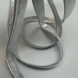 Könnte beinhalten: Nahaufnahme einer silbernen Metallborte, wahrscheinlich zum Nähen oder Basteln. Die Borte ist ein gewebter Stoff mit einer glänzenden, reflektierenden Oberfläche. Das Material ist geschlungen und gebogen, was seine Flexibilität und Textur zeigt. Die Borte ist etwa 1,3 cm breit.