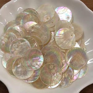 Puede incluir: Una colección de discos circulares de nácar con un acabado iridiscente. Cada pieza redonda y plana presenta un pequeño orificio centrado para enhebrar. Los discos muestran variaciones naturales en tonos blancos, crema y arcoíris.