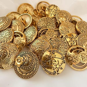 Puede incluir: Una colección de botones redondos de metal dorado con varios patrones en relieve, incluyendo motivos florales, líneas onduladas y texturas geométricas intrincadas. Cada botón tiene un ojal en la parte posterior para sujetarlo a prendas.