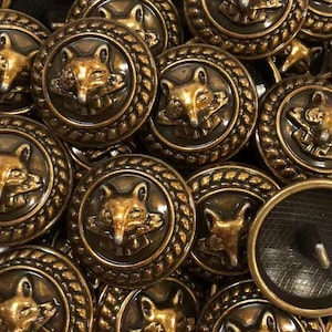 May include: A collection of round, metallic buttons featuring a raised, embossed fox head design in the center. Each button is framed by a textured, rope-like border and finished in an antique bronze tone. The back of one button shows a shank.