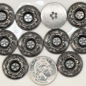 May include: A collection of silver-toned metal buttons with a floral design. Each button features a detailed flower with a central cluster of small, white accents. One button has a plain, unadorned surface with four holes. A US quarter is included for size comparison.