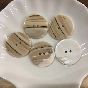 May include: Five round, two-hole buttons with a smooth, polished surface. Four buttons feature a cream and beige base with dark brown linear streaks and marbled patterns, while one button is solid white with a pearlescent, iridescent sheen.