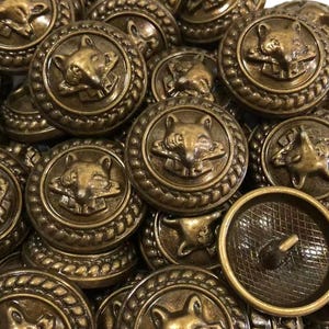 May include: A collection of round, antique brass-colored metal buttons. Each button features a raised, detailed relief of a fox head centered within a circular border with a rope-like texture. One button is flipped to show a textured back with a shank.