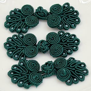 May include: Three dark green frog closures made of coiled cord and fabric loops. Each fastener features a symmetrical design with two central spiral knots and decorative fan-shaped edges accented with small, round, metallic-colored beads.