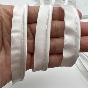 May include: Close-up of white satin bias binding, with a stitched edge. The binding is folded and held in a hand, showing the texture and sheen of the fabric. The binding is used for sewing and crafting projects.