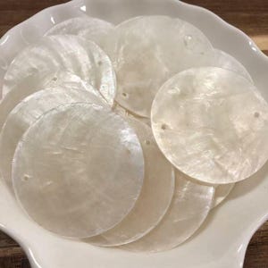 Puede incluir: Una colección de discos circulares y planos de nácar blanco. Cada disco presenta una superficie lisa e iridiscente con un brillo natural y un pequeño orificio preperforado cerca del borde, adecuado para la fabricación de joyas o manualidades.
