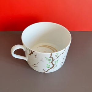 Può includere: Una tazza in ceramica bianca con una delicata illustrazione di rami d'albero marroni con piccole foglie verdi e minuscole stelle dorate. Una mezzaluna dorata è dipinta sulla base interna della tazza, dotata di un manico ricurvo.