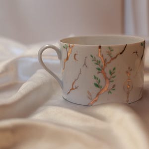 Könnte beinhalten: Eine weiße Porzellantasse mit einem zarten, handgemalten Design aus dünnen, metallisch goldenen Zweigen mit kleinen grünen Blättern. Die Tasse hat einen geschwungenen Henkel und eine glatte, glänzende Oberfläche mit goldenen Akzenten.