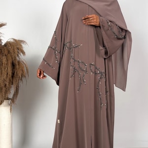 May include: A mauve-colored abaya featuring long, wide sleeves and a matching hijab. The garment is adorned with intricate dark beaded embroidery in a branch-like pattern that extends across the front bodice and down the sleeves.