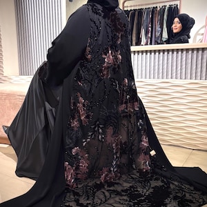 May include: A black abaya with a floral design on the back and train. The floral pattern is in shades of burgundy and black. The abaya is worn by a person in a shop.