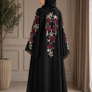 May include: A black abaya with floral embroidery in burgundy and gold. The embroidery is on the back and sleeves. The abaya is worn by a person wearing a black hijab. The abaya is floor-length and has wide sleeves.