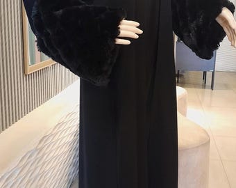 Op de afbeelding: Een lange, zwarte abaya met imitatiebont manchetten. Het kledingstuk wordt getoond op een mannequin. De abaya heeft een eenvoudig, vloeiend ontwerp en een bescheiden halslijn. De manchetten vormen een contrasterend accent.