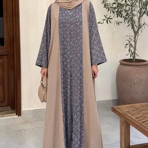 Op de afbeelding: Een lange, vloeiende beige abaya met een centraal paneel van stof met patronen in blauw- en grijstinten. De abaya heeft wijde mouwen en wordt gecombineerd met een bijpassende hoofdbedekking. Een kleine beige handtas wordt in de hand gehouden.