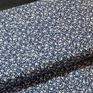 May include: Dark blue fabric covered in a close pattern of tiny, white floral motifs. The fabric is folded, revealing its texture and how it drapes. The floral design is consistently spread across the material.