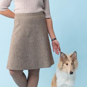 Puede incluir: Una mujer con un su&eacute;ter marr&oacute;n claro y una falda marr&oacute;n est&aacute; de pie con un perro Collie marr&oacute;n y blanco. La mujer sonr&iacute;e y mira a la c&aacute;mara. El perro est&aacute; sentado y mirando a la c&aacute;mara.