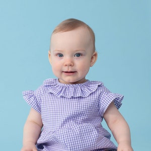 May include: A baby wearing a purple and white gingham romper with a frilled collar. The baby is sitting on a light blue background.