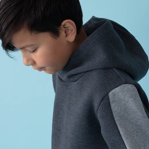 Könnte beinhalten: Ein dunkelgrauer Kapuzenpullover mit einem grauen Patch am linken Ärmel. Der Pullover ist aus einem weichen, bequemen Stoff und hat eine lockere Passform.