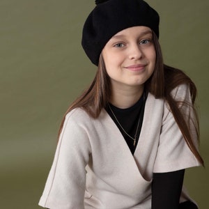 May include: A young person wearing a black beret with a pom-pom on top, a beige sweater, and black trousers. They are sitting on a wooden stool and smiling at the camera.