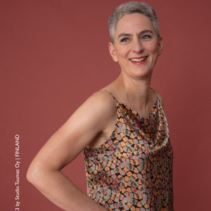 May include: A woman wearing a sleeveless top with a cowl neck and a geometric pattern in shades of orange, brown, green, and red. The top is made of a silky fabric.