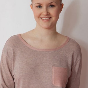 May include: A woman wearing a long-sleeved, light pink shirt with a small, light pink pocket on the left side. The shirt is made of a soft, textured fabric.