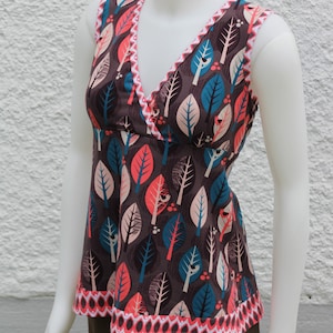 May include: A sleeveless brown top with a V-neckline and a pink and blue leaf pattern. The top has a pink and white scalloped trim around the neckline and hem. The text "OTTOBRE design® © STUDIO TUUMAT OY" is printed at the bottom of the image.
