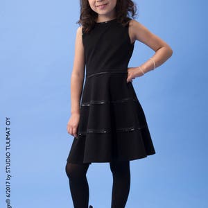 May include: A young girl wearing a black sleeveless dress with a layered skirt and black tights. She is wearing black patent leather boots with laces. The girl is standing on a light blue background.