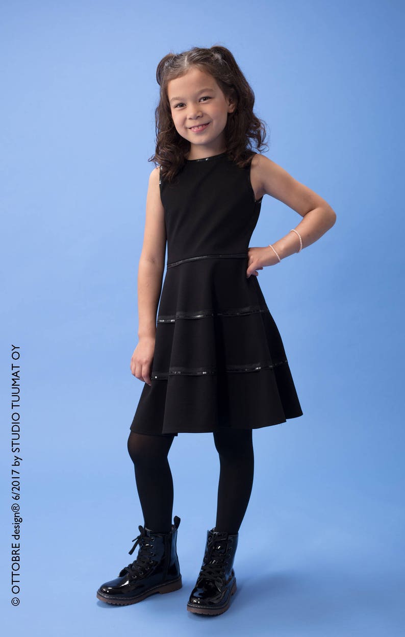 May include: A young girl wearing a black sleeveless dress with a layered skirt and black tights. She is wearing black patent leather boots with laces. The girl is standing on a light blue background.