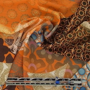 May include: A close-up of a multi-colored fabric with a variety of geometric patterns. The fabric is orange, brown, and blue with a white background. The fabric is folded in half and the ruler is visible at the bottom of the image.