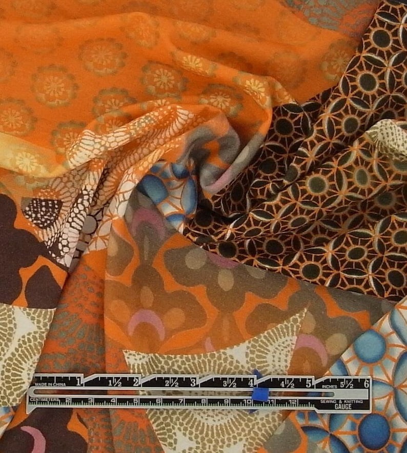 May include: A close-up of a multi-colored fabric with a variety of geometric patterns. The fabric is orange, brown, and blue with a white background. The fabric is folded in half and the ruler is visible at the bottom of the image.