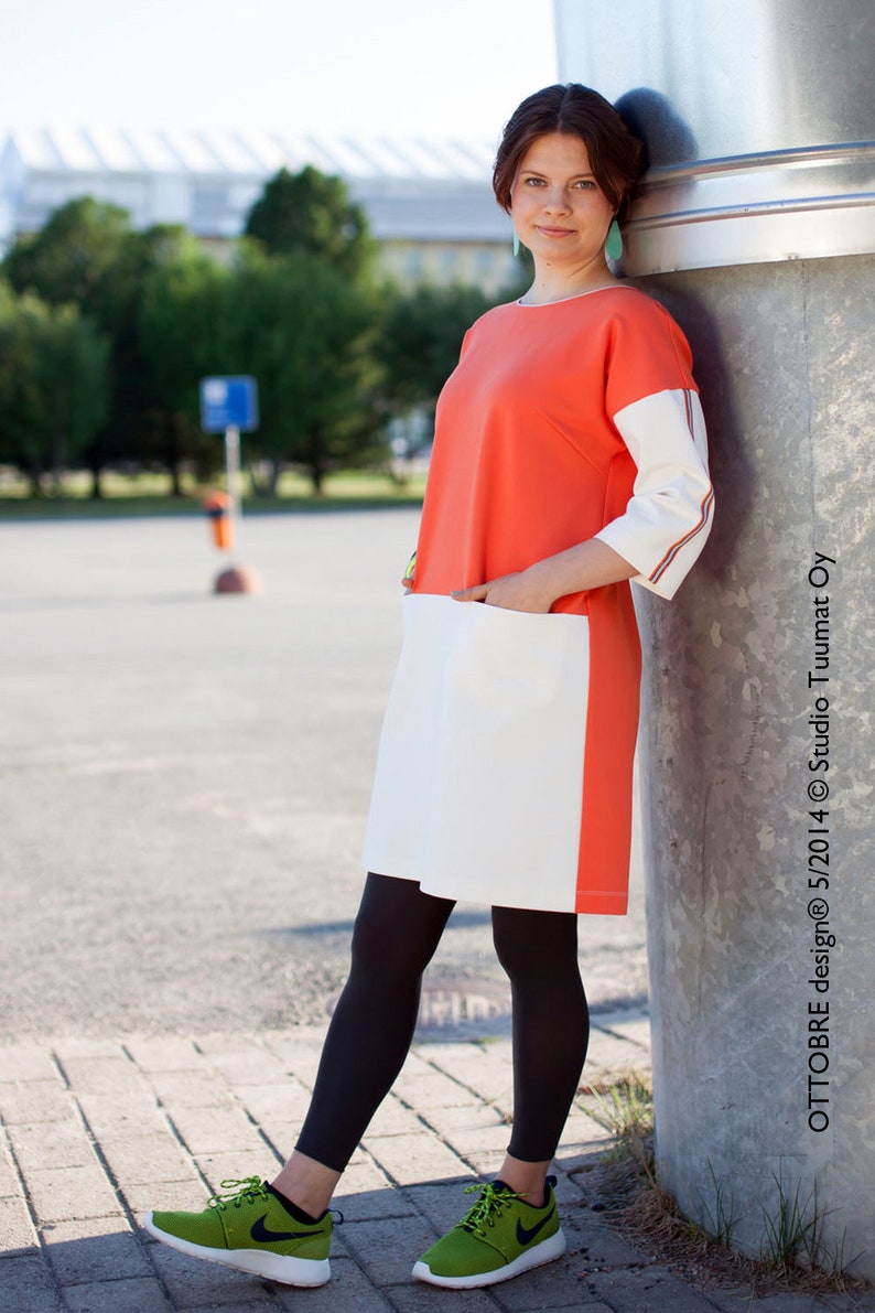 May include: A woman wearing a coral and white dress with black leggings and green sneakers. She is standing next to a silver pole. The dress has a white pocket on the front and a stripe of orange, white, and blue on the sleeve. The text "OTTOBRE design&reg; 5/2014 &copy; Studio Tuumat OY" is on the pole.