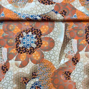 May include: A fabric with a repeating pattern of orange, brown, and blue flowers. The flowers are made up of smaller circles and swirls. The background is white with a subtle pattern of small circles.
