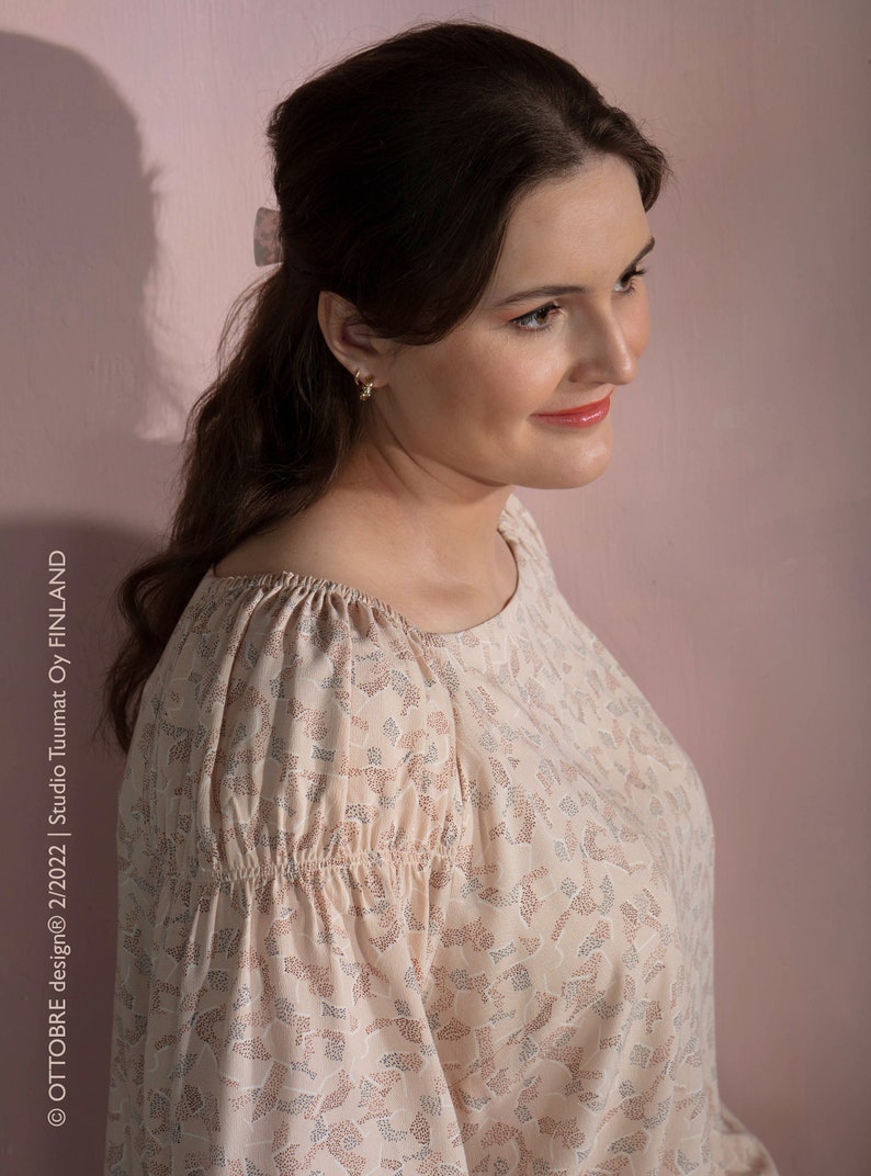 May include: A woman wearing a light pink blouse with a small floral pattern. The blouse has a gathered neckline and long sleeves.