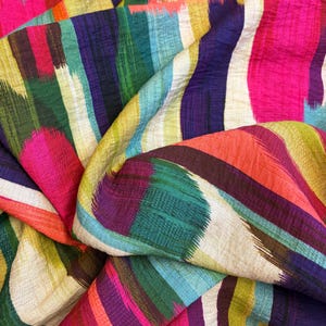 May include: A close-up of a colourful fabric with a vibrant, abstract design. The fabric features vertical brushstrokes in shades of pink, purple, green, blue, orange, and yellow. The fabric is folded, creating a textured and dynamic visual.