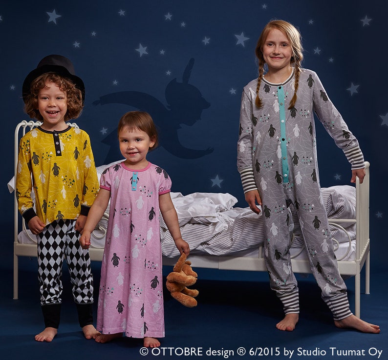 May include: Three children in pyjamas stand in front of a bed against a dark blue backdrop with stars. One child wears a yellow long-sleeved shirt with black and white diamond-patterned trousers. Another wears a pink nightdress. The third child wears a grey jumpsuit.