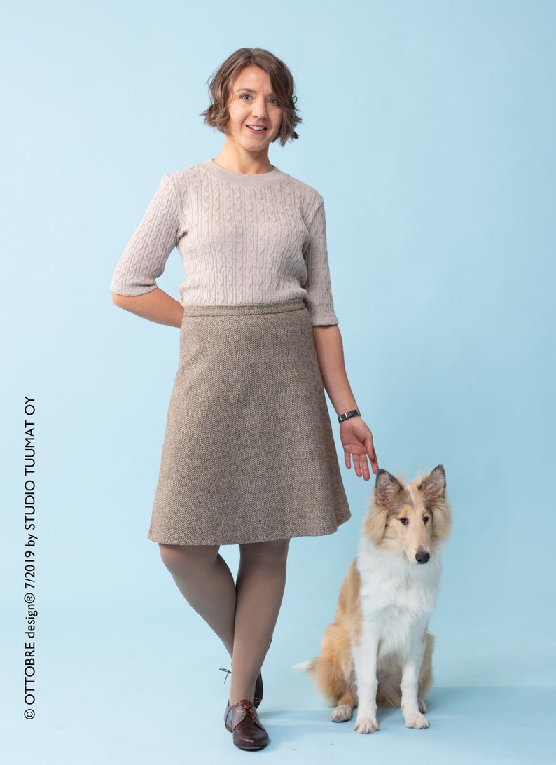 Puede incluir: Una mujer con un su&eacute;ter marr&oacute;n claro y una falda marr&oacute;n est&aacute; de pie con un perro Collie marr&oacute;n y blanco. La mujer sonr&iacute;e y mira a la c&aacute;mara. El perro est&aacute; sentado y mirando a la c&aacute;mara.