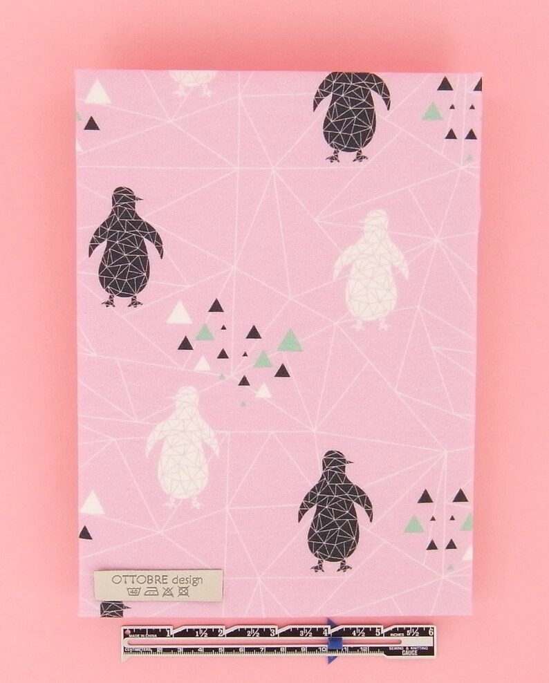May include: Pink fabric featuring a geometric design of penguins in black and white, with small triangles in black, white, and green. The fabric has a label that reads "OTTOBRE design". A ruler is at the bottom.