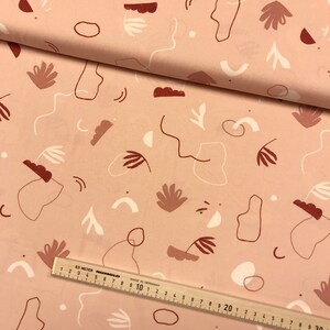 May include: A pink fabric with a white and brown abstract pattern. The pattern includes circles, lines, and shapes. The fabric is folded in half and a ruler is visible at the bottom of the image.
