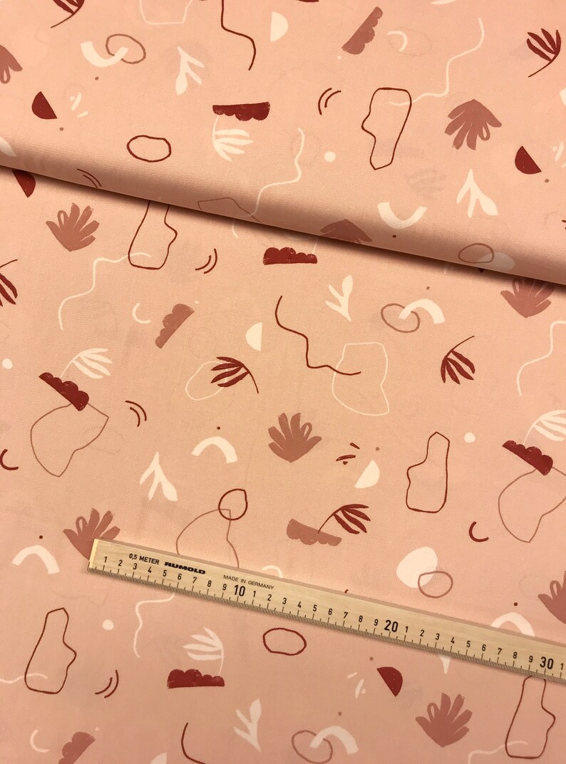 May include: A pink fabric with a white and brown abstract pattern. The pattern includes circles, lines, and shapes. The fabric is folded in half and a ruler is visible at the bottom of the image.