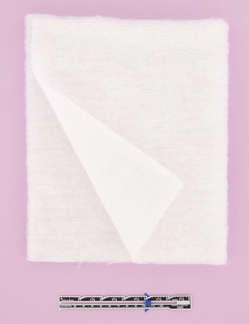 Puede incluir: Una pieza blanca rectangular de guata doblada por la mitad. La guata mide aproximadamente 15 cm de ancho y 15 cm de largo.