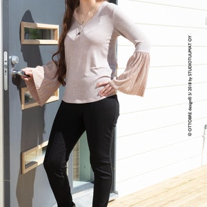 May include: A woman wearing a light brown long-sleeved shirt with bell sleeves, black pants, and black ankle boots. She is standing in front of a gray door with glass panels and a white wooden wall.