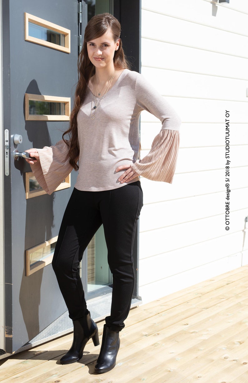 May include: A woman wearing a light brown long-sleeved shirt with bell sleeves, black pants, and black ankle boots. She is standing in front of a gray door with glass panels and a white wooden wall.
