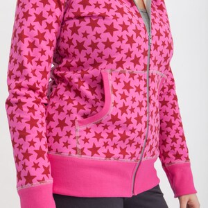 Puede incluir: Una sudadera con capucha rosa con cremallera y un estampado de estrellas rojas. La sudadera tiene capucha y un bolsillo delantero.