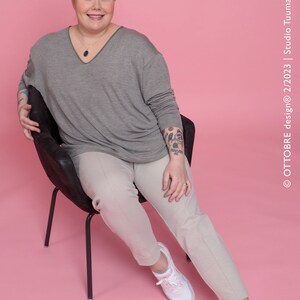 May include: A woman wearing a gray long-sleeved shirt, beige pants, and white sneakers with pink accents. She is sitting on a black chair against a pink background. She has a tattoo on her arm.