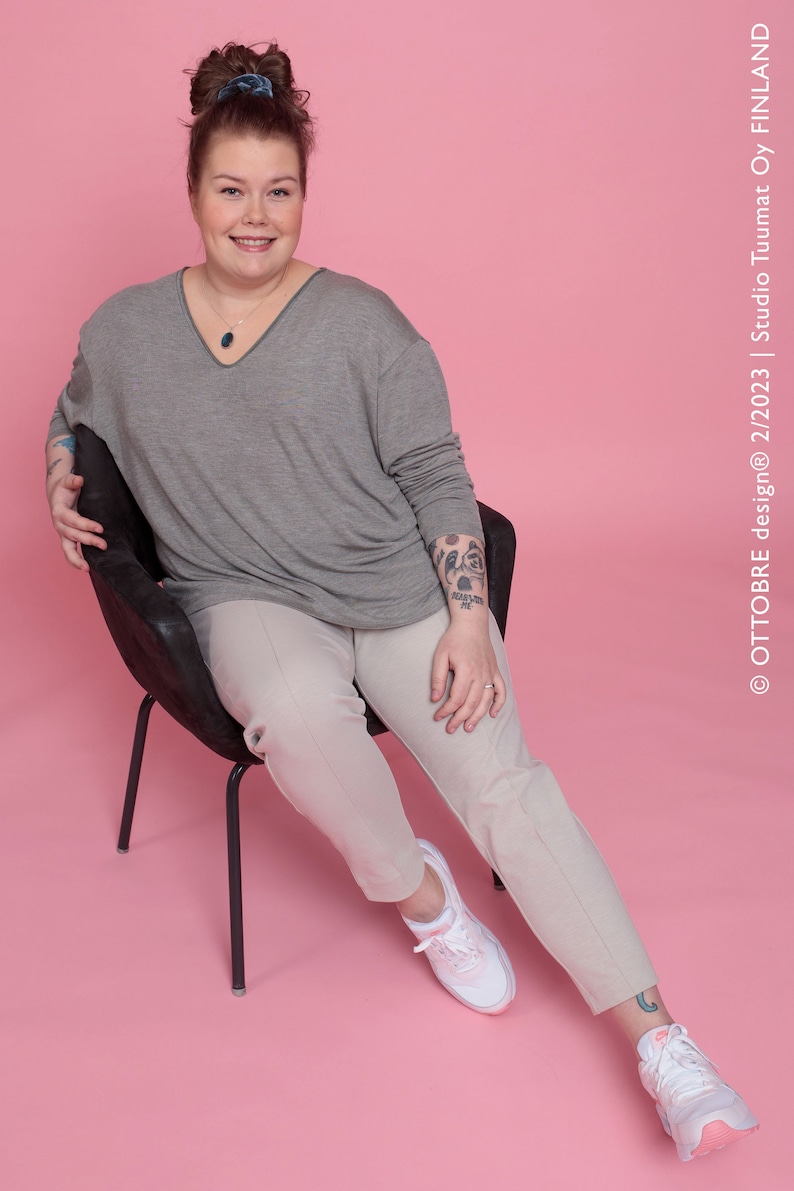 May include: A woman wearing a gray long-sleeved shirt, beige pants, and white sneakers with pink accents. She is sitting on a black chair against a pink background. She has a tattoo on her arm.