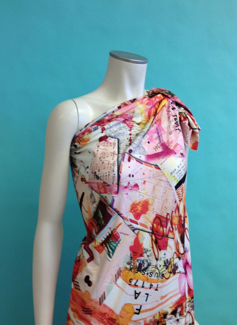 May include: One-shoulder dress with a colorful abstract print featuring musical notes, letters, and splashes of color. The dress is made of a soft, flowing fabric.